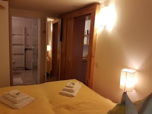 Una habitación de hotel con una cama, una lámpara de noche, una puerta que da a un baño y una mesita con una toalla.
