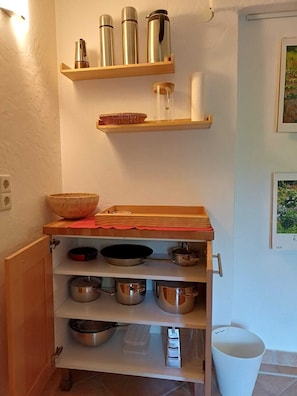 Una cocina con estantes de madera, ollas de acero inoxidable y un cubo de basura blanco.