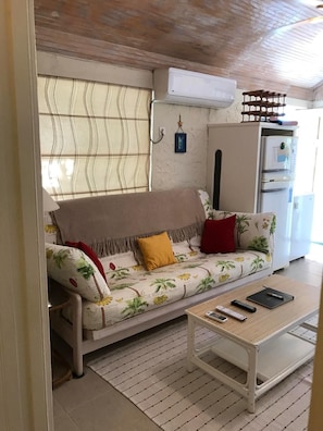 A living room with a sofa, a coffee table, a refrigerator, and a window with blinds.