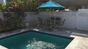 Ein rechteckiger Pool mit Metallleiter, umgeben von einem weißen Zaun, und ein Tisch mit Stühlen unter einem blauen Sonnenschirm.
