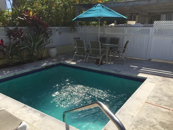 Una piscina rectangular con una escalera de metal, rodeada por una cerca blanca, y una mesa con sillas bajo una sombrilla azul.