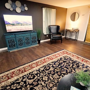A living room with a flat-screen TV, a dark leather chair, a blue cabinet, a patterned rug, and a small table with a 'HELLO' sign.