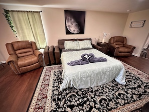 A bedroom with a bed, two armchairs, a nightstand, a wall-mounted picture, and a decorative plant.