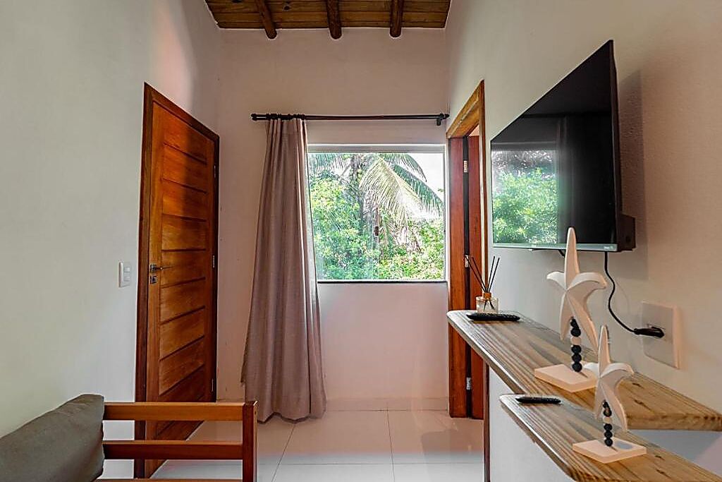 A room with a wooden door, a window with a view of greenery, a flat-screen TV mounted on the wall, and a wooden bench.