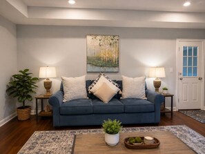 Ein Wohnzimmer mit einem blauen Sofa, zwei Lampen, einem Gemälde, einer Topfpflanze und einem Couchtisch mit Dekoration.