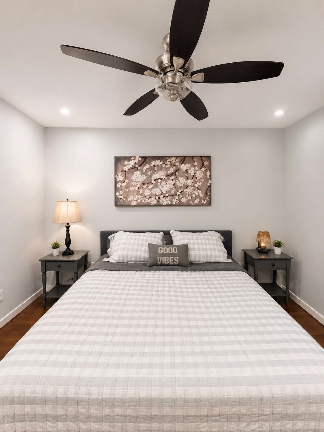 A bedroom with a bed, a ceiling fan, a wall-mounted artwork, bedside tables with lamps, and a pillow with text.
