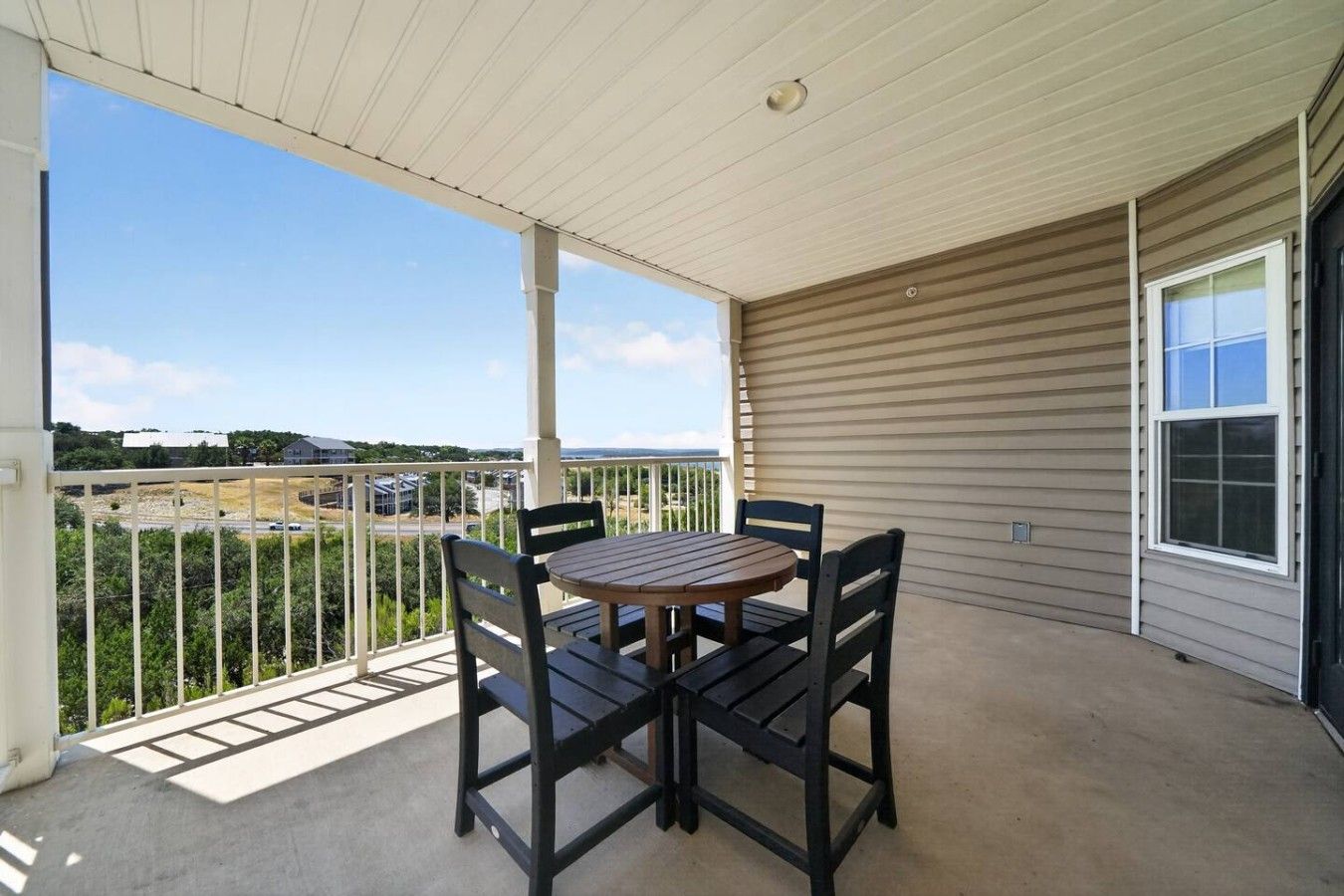 A covered balcony with a table and chairs, a view of the landscape, and a building with siding.