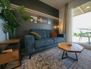 Een moderne woonkamer met een blauwe bank, een houten salontafel, een gekleurd vloerkleed en een grote plant.