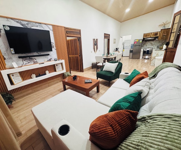 A living room with a TV, sofa, coffee table, and kitchen in the background.