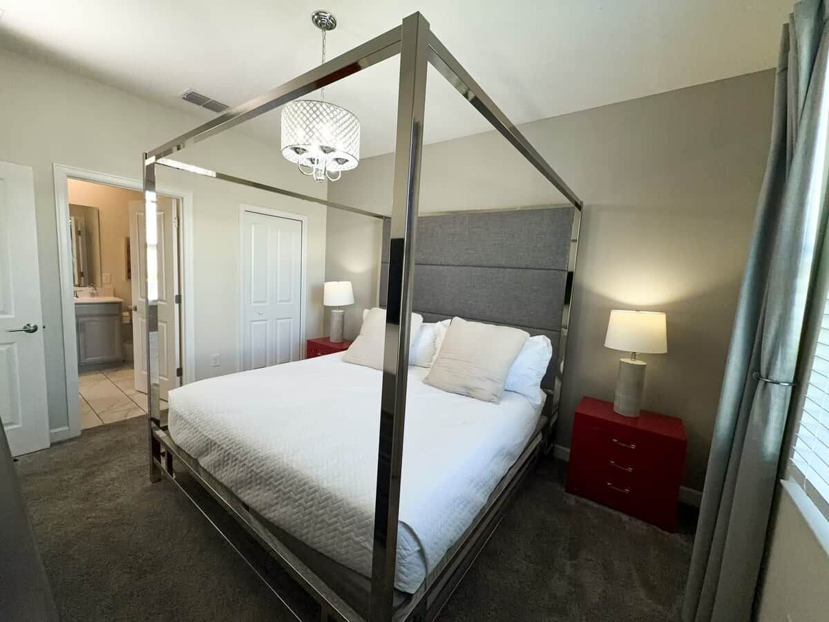 A four-poster bed with a grey headboard, two bedside tables with lamps, and a door leading to a bathroom.