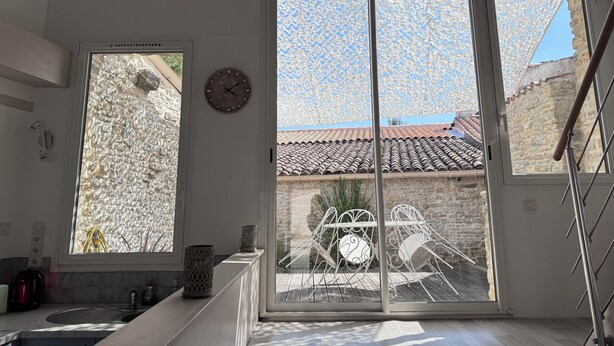 A room with a large window, a clock, and a view of a tiled roof and stone wall. - Maison de la Sau (Saint-Georges-d'Oléron)