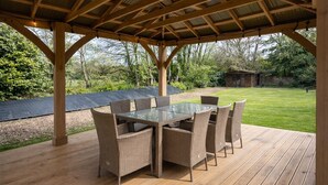 A covered outdoor dining area with a glass table and wicker chairs, surrounded by a wooden deck and a grassy lawn.