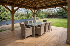 A covered outdoor dining area with a glass table and wicker chairs, surrounded by a wooden deck and a grassy lawn. - Elegy House (Slough)