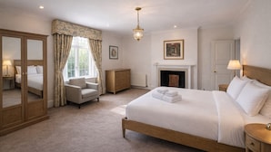 A hotel bedroom with a large bed, a sitting area with a chair, a dresser, a fireplace, and a mirror.