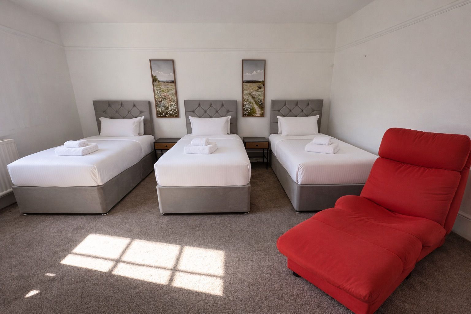 Quarto de hotel com três camas, uma cadeira vermelha e duas pinturas penduradas na parede.