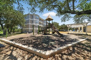 A playground with a slide and climbing structures, surrounded by trees and a building in the background. - Relaxing 2BR w/ AC, WiFi · Canyon Lake #420 (Canyon Lake)