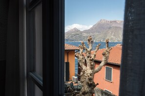 A coastal view through a window, featuring a tree, buildings with red roofs, and mountains in the background. - Varenna Boulevard: Spacious Apartment in the City Center (Varenna)