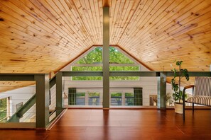 Una habitación amplia con techo de madera inclinado, una escalera y un gran ventanal con vistas a la vegetación.