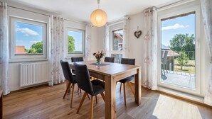 Een eetgedeelte met een houten tafel, zwarte stoelen, een hanglamp en grote ramen met gordijnen.