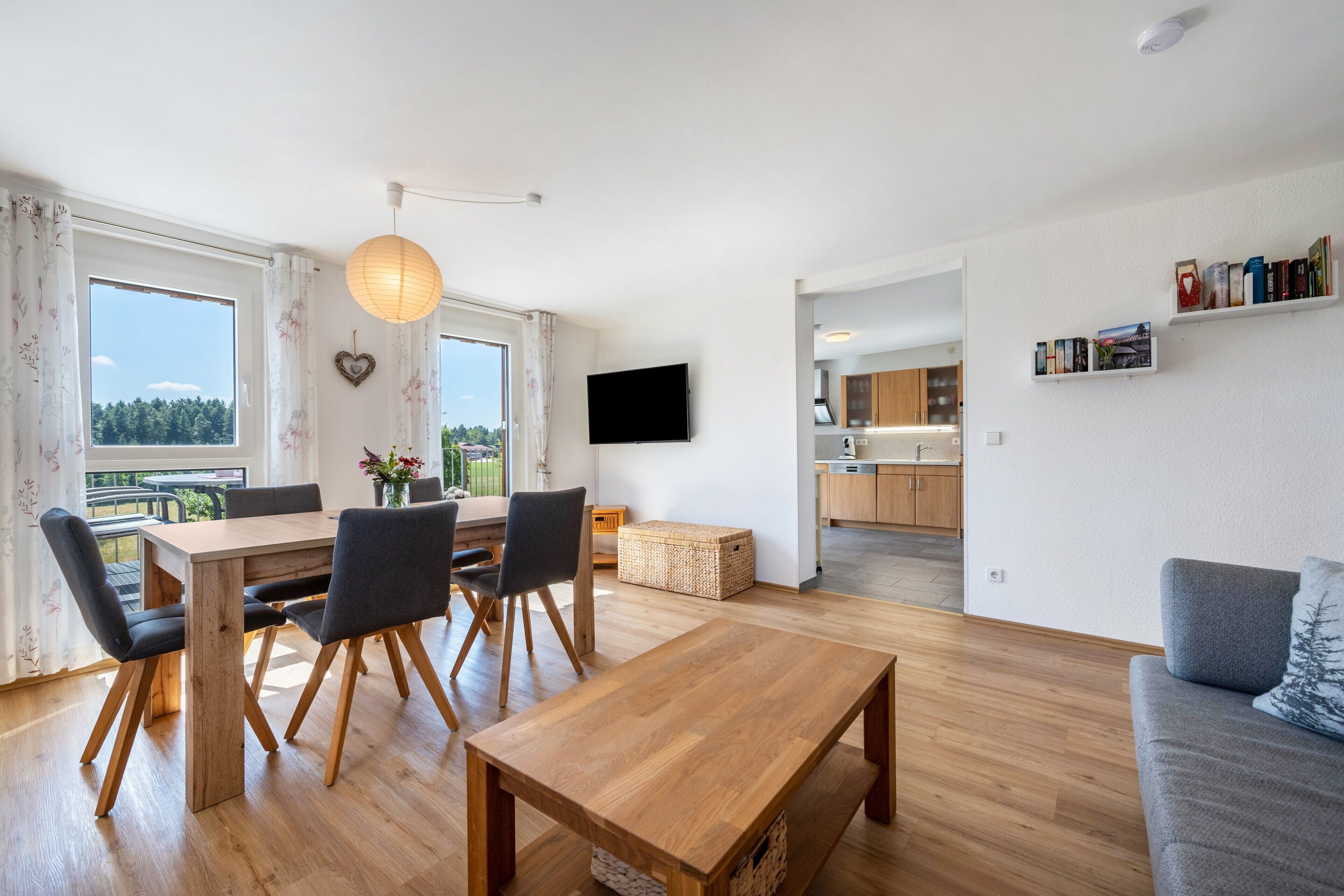 A modern living space with a dining area, a sofa, a TV, and a kitchen visible through an open door.