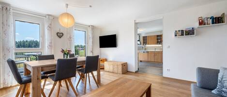Un espace de vie moderne comprenant un coin repas, un canapé, un téléviseur et une cuisine visible à travers une porte ouverte.