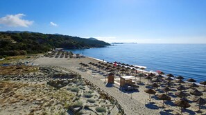 A beach with umbrellas and sun loungers, a fenced area with plants, and a clear view of the sea and distant hills.