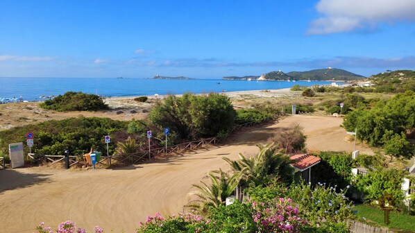 A coastal view with a dirt path leading to a beach, surrounded by greenery and a clear blue sky.