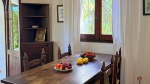 Een eetgedeelte met een houten tafel, stoelen, een fruitschaal, een decor kast en een raam met uitzicht op groen.