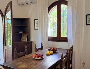 A dining area with a wooden table, chairs, a fruit bowl, a decorative cabinet, and a window with a view of greenery.