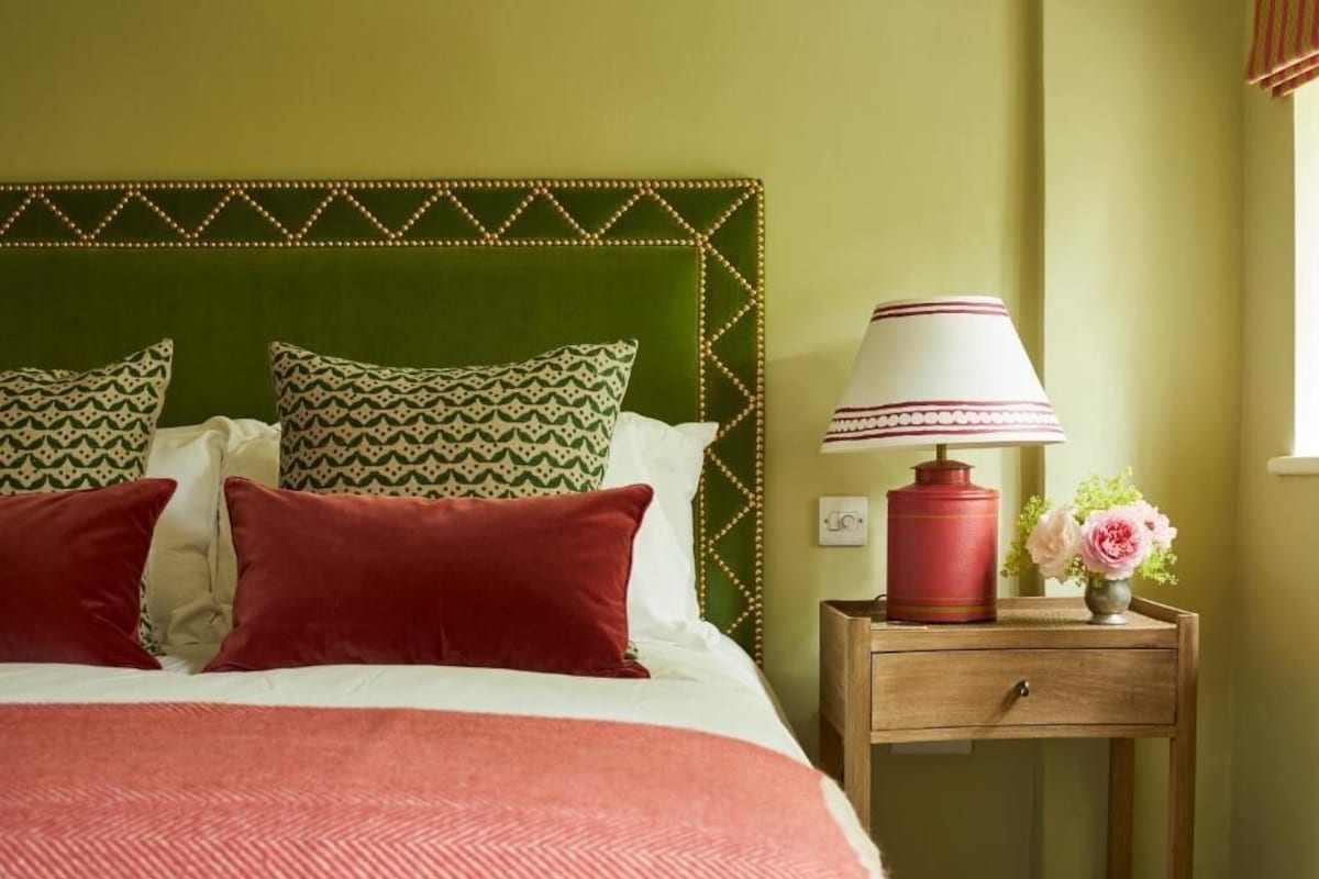 A bed with patterned pillows, a red lamp, a small table with a vase of flowers, and a green headboard.