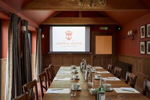 A conference room with a long table set for a meeting, a screen displaying 'CROWN & ANCHOR' branding, wooden paneling, and framed pictures on the walls.