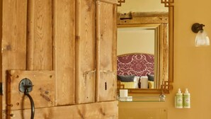 A wooden wardrobe with a black handle, a mirror with a decorative frame, a sink with two faucets, and a shelf with toiletries. - Crown and Anchor Ham (Marlborough)