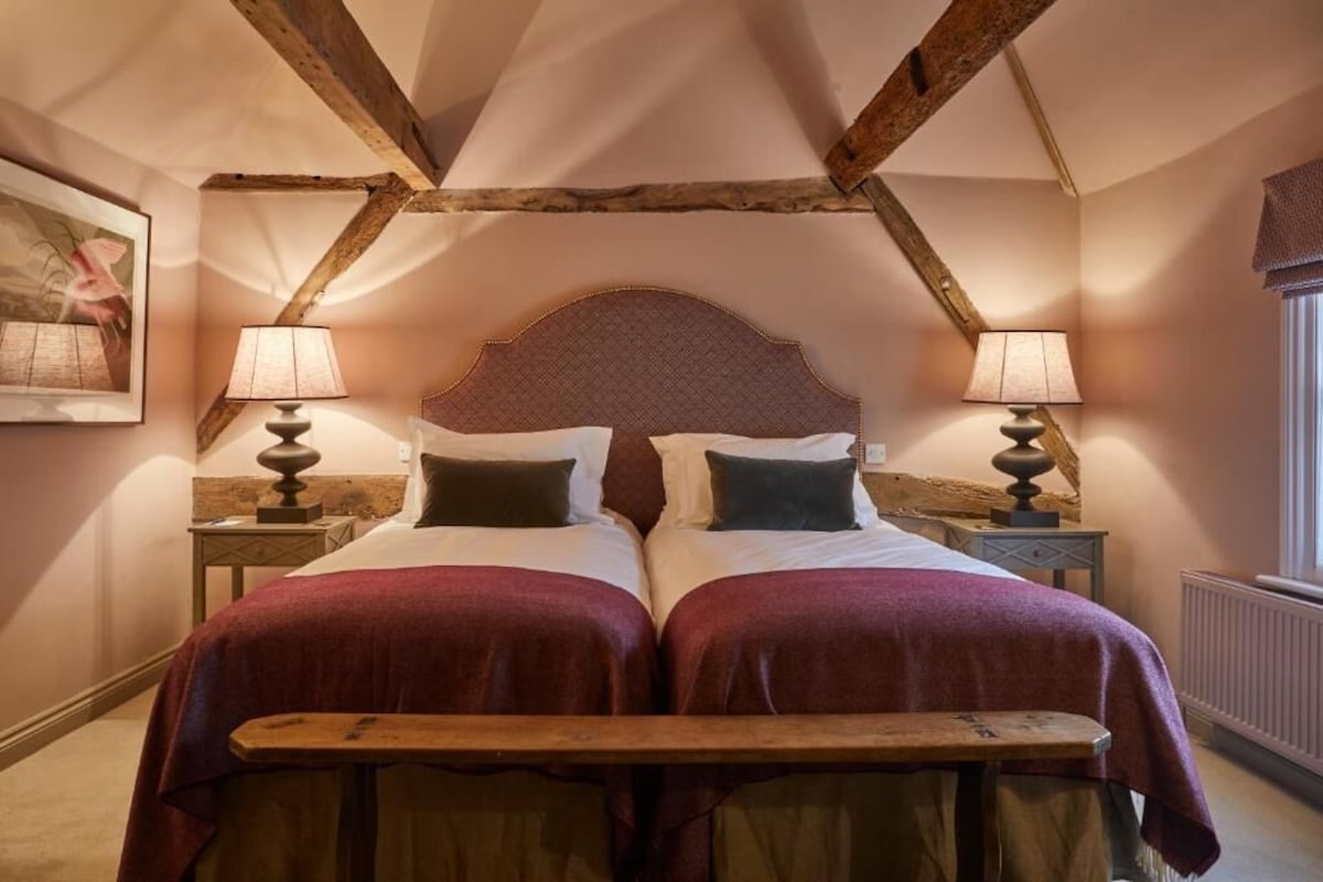 A room with a high ceiling, exposed wooden beams, two beds with red bedding, bedside lamps, and a framed picture on the wall.