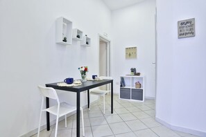 Une petite pièce propre, avec une table noire, des chaises blanches, une étagère blanche ornée d’objets décoratifs et une porte donnant sur une autre pièce.