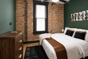 Ein Schlafzimmer mit Backsteinwand, einem Fenster mit Jalousien, einem Bett mit weißer Bettwäsche und braunen Akzenten, einer Holzkommode und einem schwarz gemusterten Teppich.