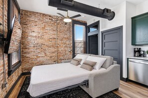 A compact room with a bed, a brick wall, a ceiling fan, and a kitchenette area.