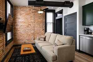 A living room with a brick wall, a sofa, a coffee table, and a kitchen area with a refrigerator.