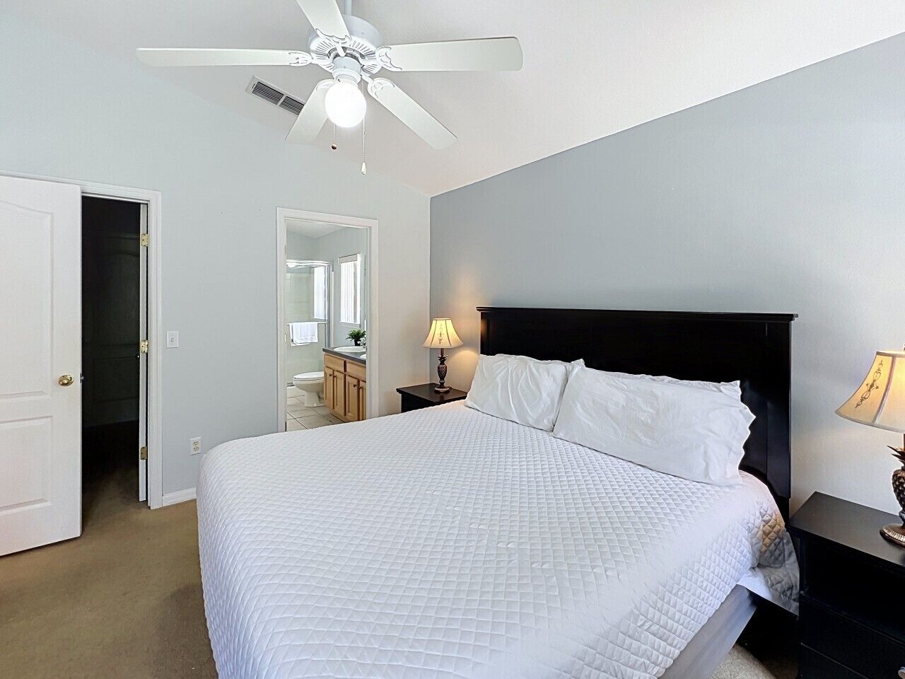 Une chambre à coucher avec un lit, un ventilateur de plafond, une table de chevet avec une lampe, une salle de bain visible à travers une porte ouverte, et une porte.