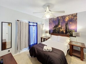 Ein Hotelzimmer mit Bett, Deckenventilator, Nachttisch, Lampe, Fenster mit Vorhängen, Wandspiegel und einem großen Poster.