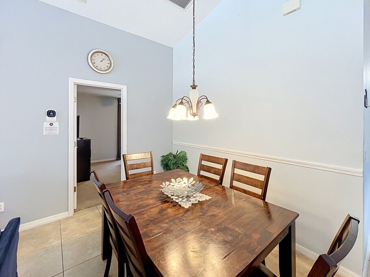 Un comedor con una mesa de madera, cuatro sillas, una lámpara de araña, un reloj y una puerta que da a otra habitación.
