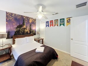 Ein Schlafzimmer mit einem Bett, einem Deckenventilator, einem Nachttisch, einer Lampe, einer Wand mit Flaggen und einer Tür.