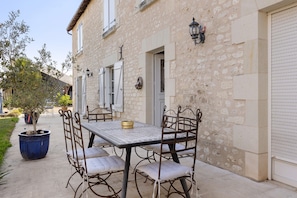 Un bâtiment en pierre avec une table et des chaises sur une terrasse, un pot de fleurs et une porte-volet fermée.