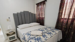 Un dormitorio con cama, mesita de noche, lámpara y ventana con cortinas.
