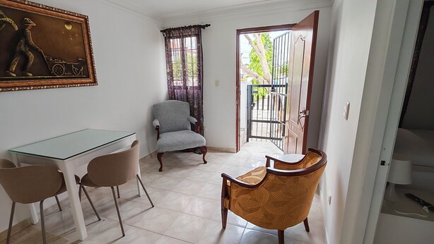 Un comedor con una mesa de vidrio y sillas, un sillón gris, un sillón estampado y una lámpara en el techo.