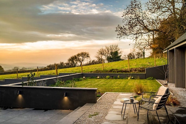 An outdoor patio with a table and chairs, a garden view, and a modern building in the background.