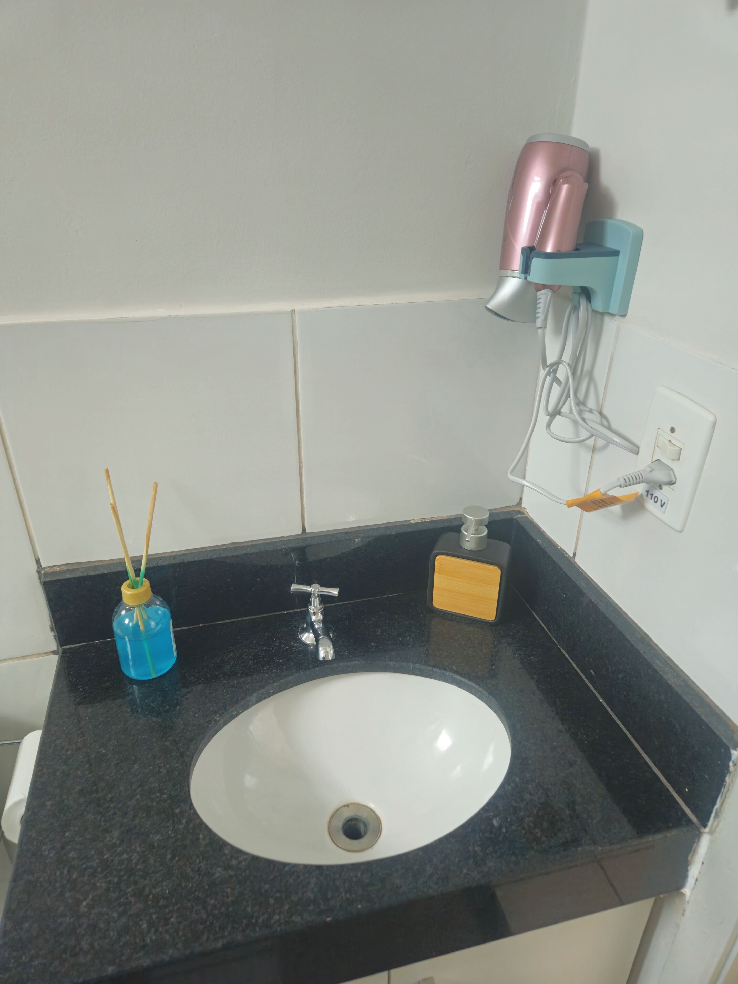 Un lavabo con encimera negra, un lavabo blanco, una botella azul, un dispensador de jabón amarillo y un secador de pelo rosa sobre una repisa de pared.