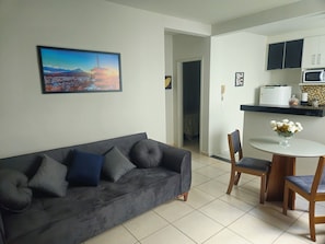 Un salon comprenant un canapé, des fauteuils, une table à manger, un coin cuisine avec des placards et un four à micro-ondes, ainsi qu’un tableau encadré accroché au mur.