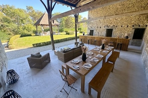 An outdoor dining area with a stone wall, a wooden table set for six, and a stone fireplace.