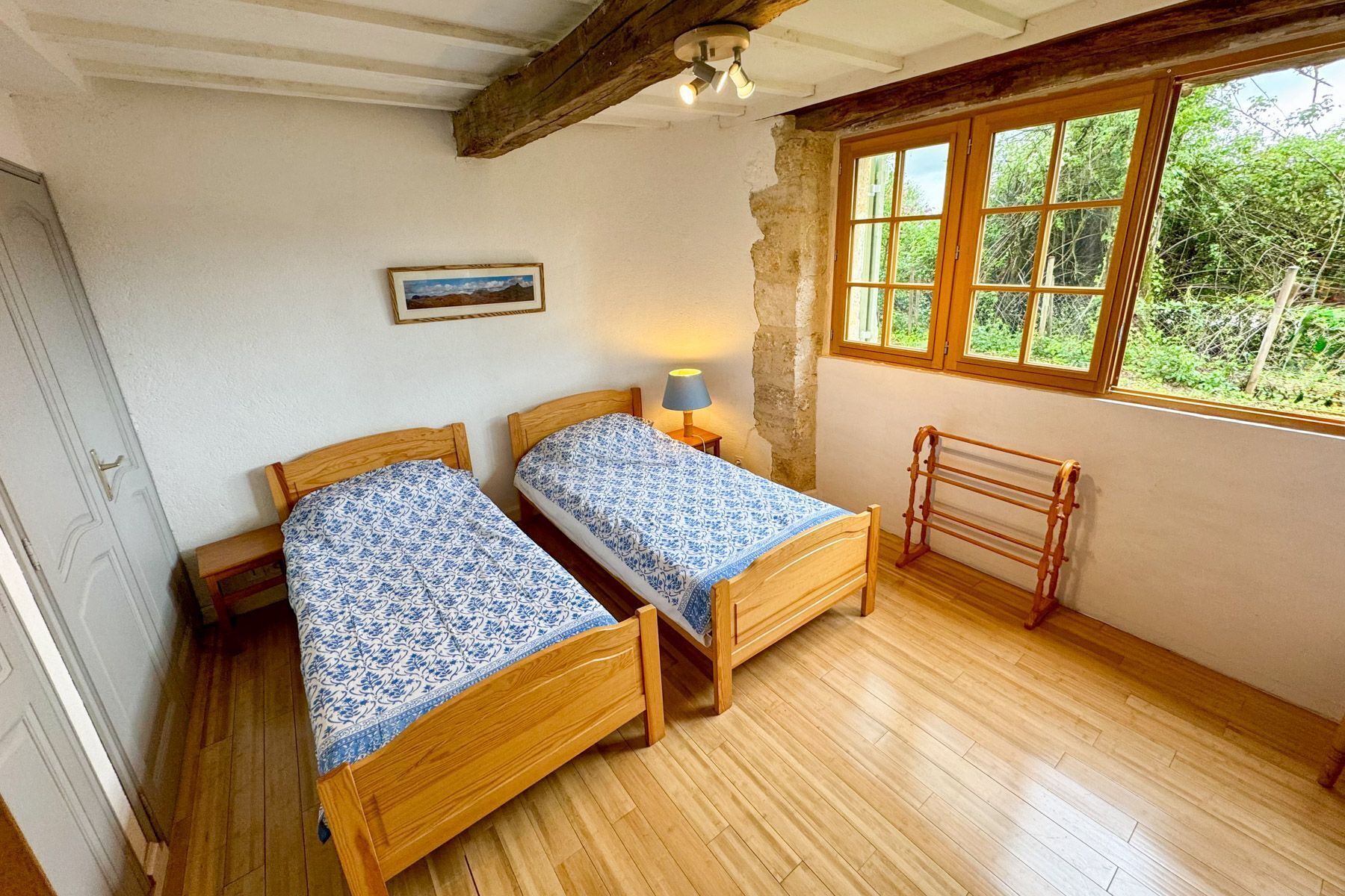 A bedroom with two wooden beds, a wooden drying rack, a single lamp, and a window with a view of greenery.