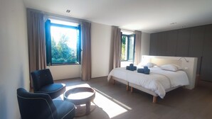 Ein modernes Hotelzimmer mit einem Bett, zwei Sessel, einem kleinen runden Tisch und großen Fenstern mit Vorhängen.